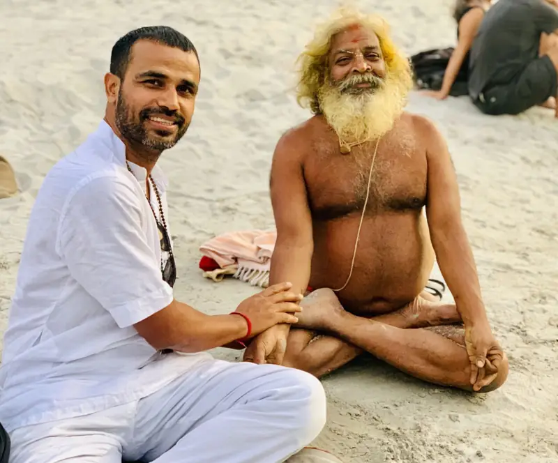 A teacher in white sitting beside a smiling sadhu on the sand