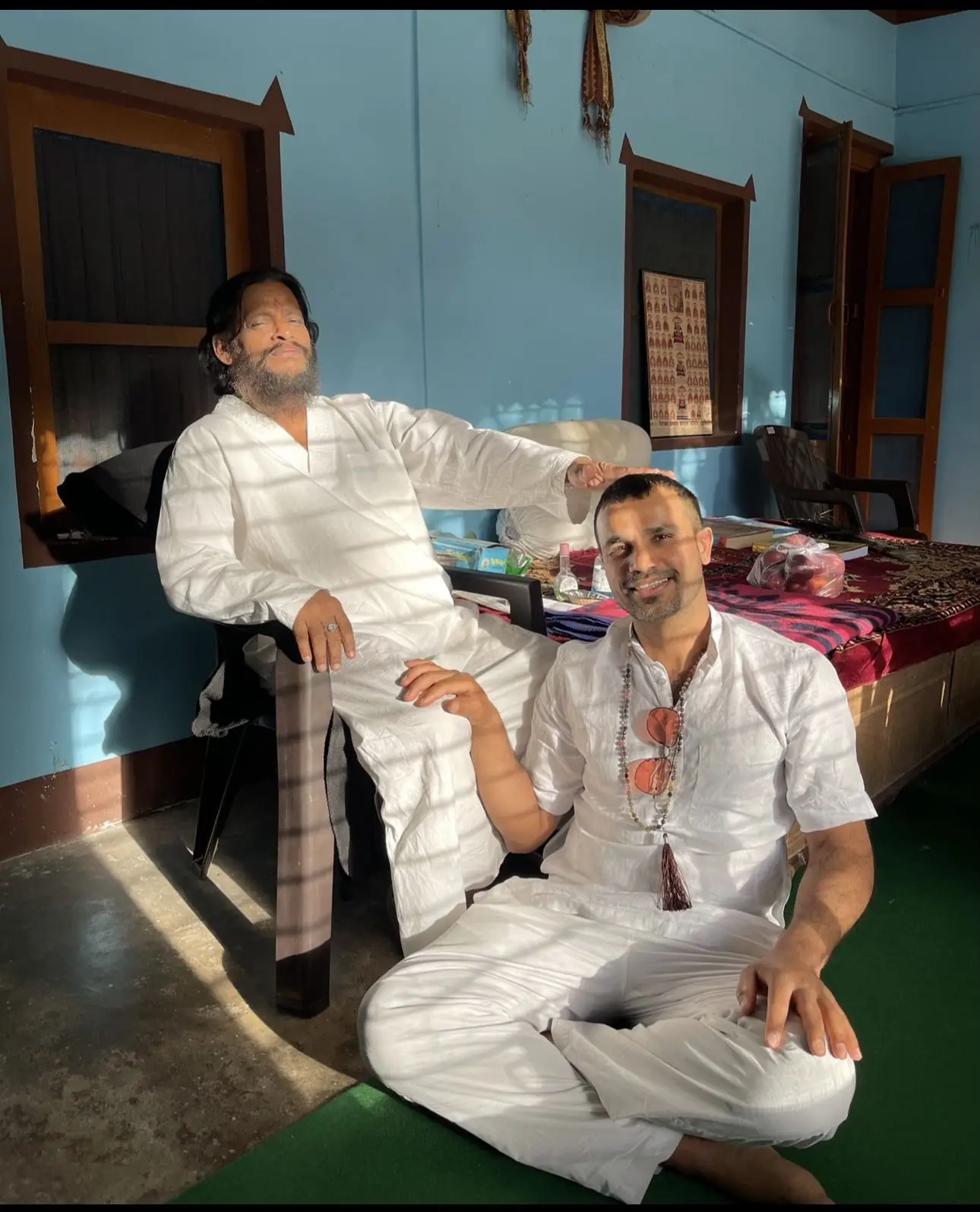 A teacher sitting at the feet of his own guru in a sunlit room in India