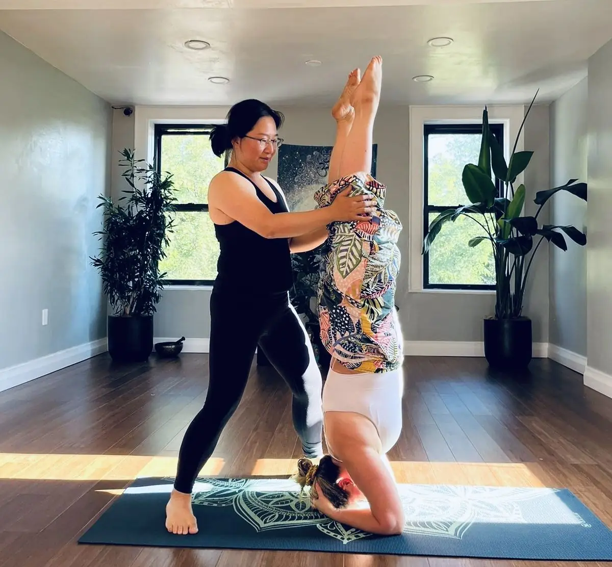 A teacher supporting a student in headstand by the studio windows